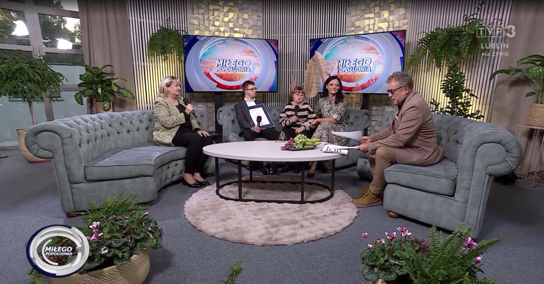 grupa pięciu osób siedzących w studiu telewizyjnym. Siedzą na szarych kanapach, ustawionych wokół okrągłego stolika z owocami. Część osób trzyma mikrofony, biorą udział w rozmowie na żywo. W tle są ekrany z napisem „Miłego Popołudnia” oraz kilka roślin, które dodają przytulności scenie.