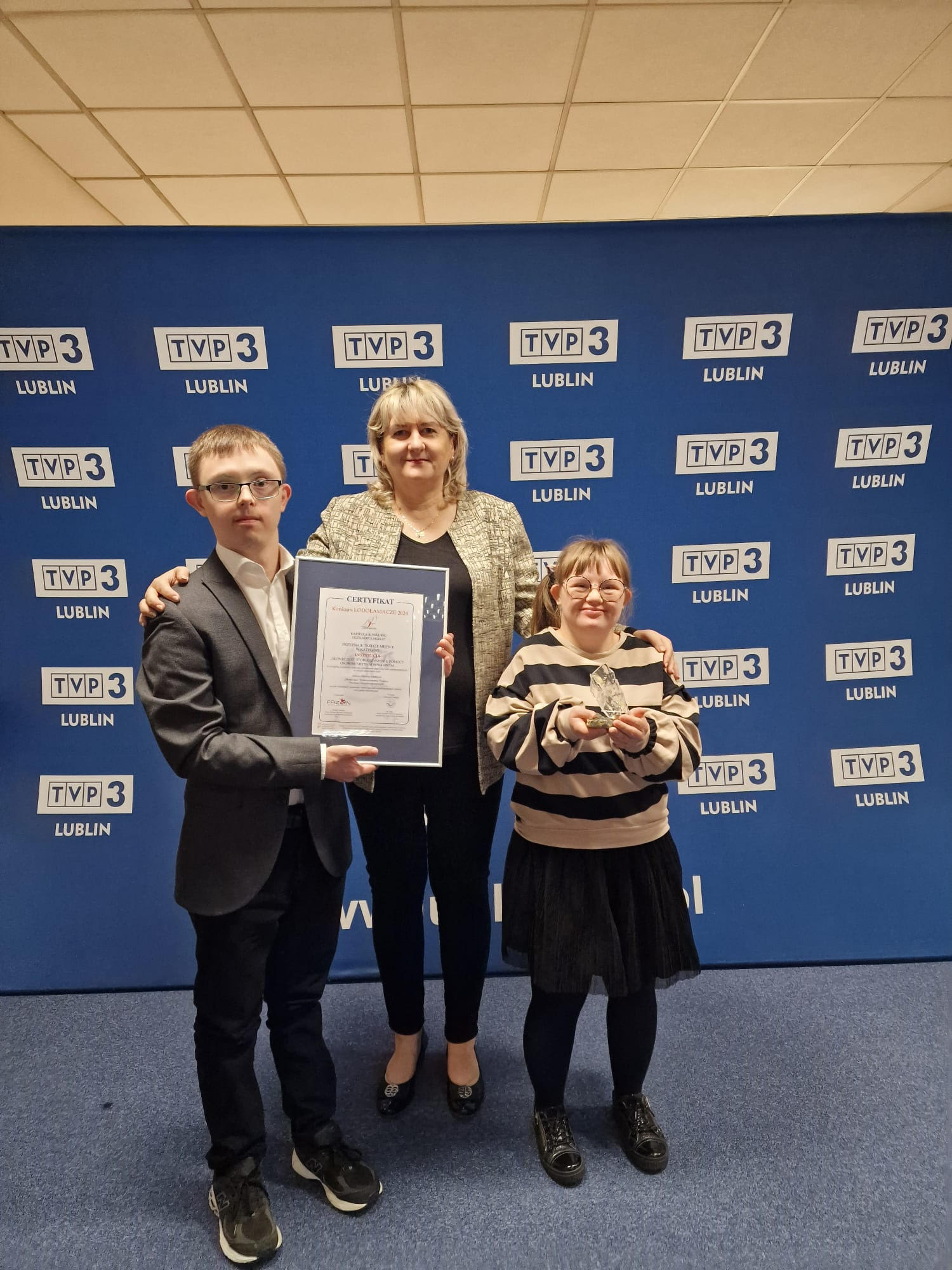 3 osoby stoją na tle baneru z logo TVP3Lublin. W środku stoi pani Prezes Dorota Fornalska, z dwóch stron chłopiec i dziewczynka. Chłopiec trzyma dyplom oprawiony w ramkę
