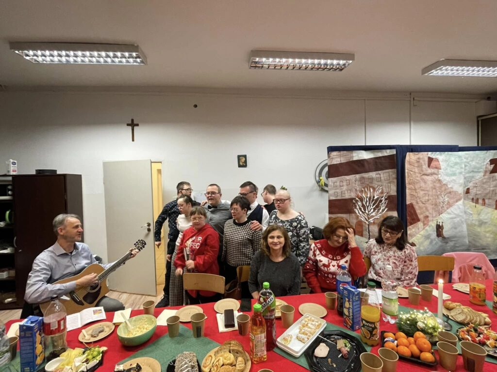 Grupa osób stoi i śpiewa. Obok jeden pan siedzi na krześle i gra na gitarze