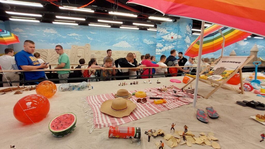 Uczestnicy wycieczki obserwujący makietę kolejową Kolejkowo, przedstawiającą scenę plaży z piaskiem, parasolem i miniaturowymi elementami.