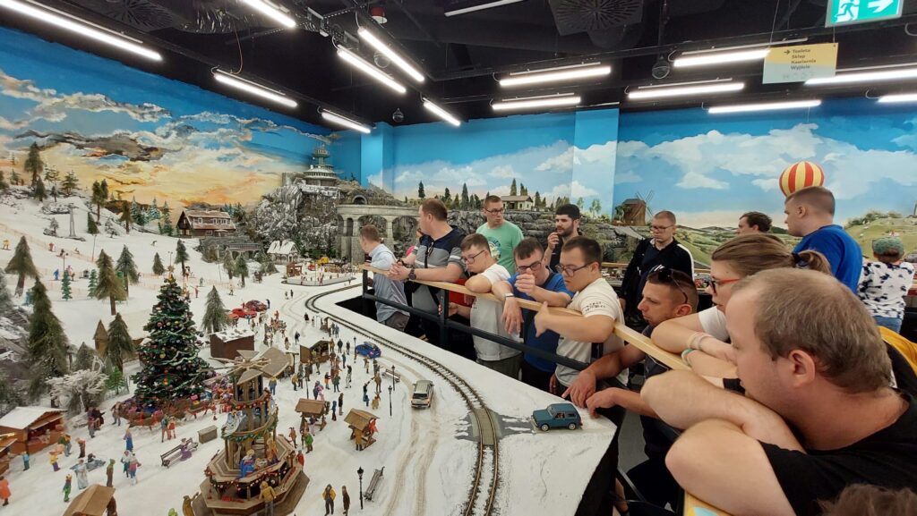 Podopieczni stowarzyszenia Słoneczko oglądający z bliska dużą, zimową makietę kolejową z ośnieżonymi górami, torami i miniaturowymi domkami.