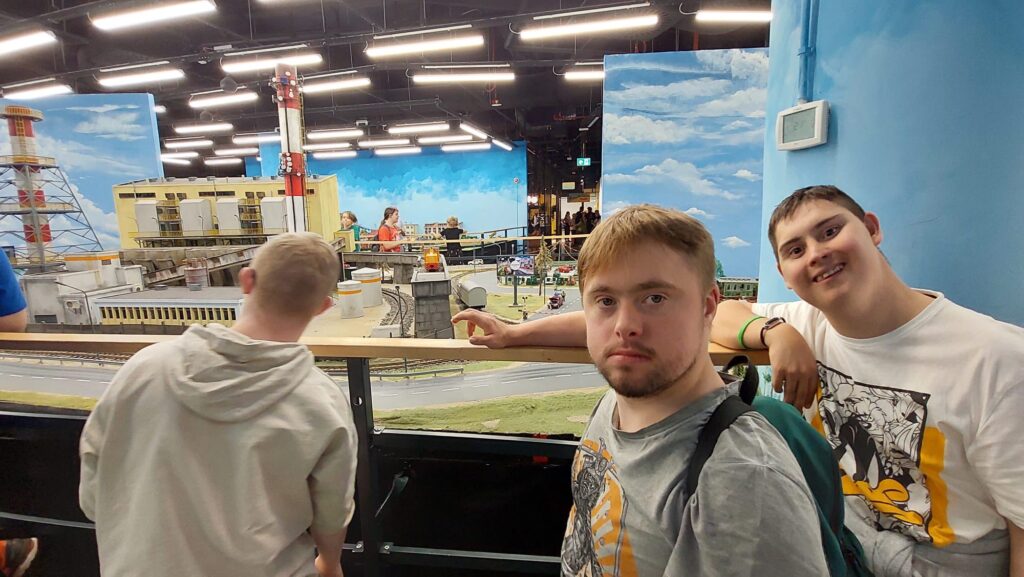 Dwóch podopiecznych uśmiecha się do kamery na tle fragmentu dużej makiety kolejowej w pomieszczeniu wystawowym Kolejkowo.