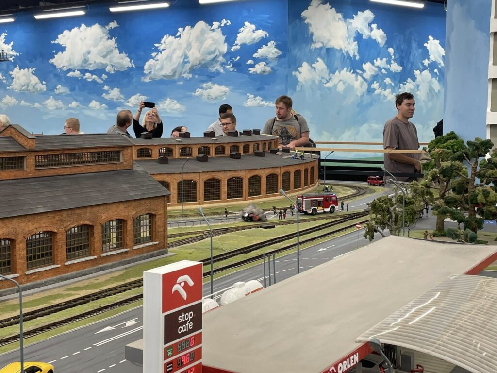Grupa osób obserwująca realistyczną makietę kolejową z miniaturową lokomotywownią, stacją benzynową Orlen i wozem strażackim.
