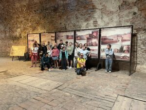 Grupa osób na tle wielkoformatowych plansz historycznych w ceglanym, sklepionym wnętrzu fortyfikacji lub podziemi.