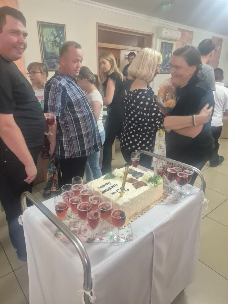 Uczestnicy spotkania lub przyjęcia w pomieszczeniu, wokół stolika z tortem z napisem oraz napojami