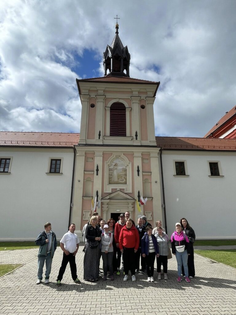 Grupa osób pozująca na tle wysokiego, zabytkowego kościoła lub klasztoru z charakterystyczną wieżą i dzwonnicą.