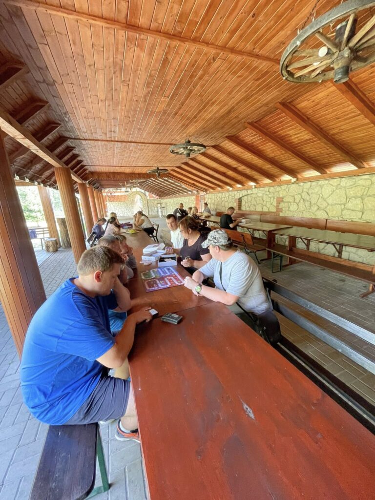 Grupa osób siedząca przy długich drewnianych stołach pod zadaszoną wiatą, podczas  gier planszowych.