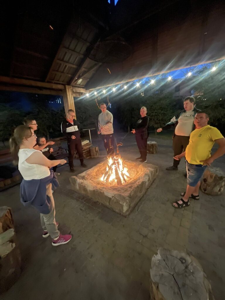 Grupa osób stojących wokół dużego, murowanego paleniska na świeżym powietrzu podczas wieczornego ogniska.