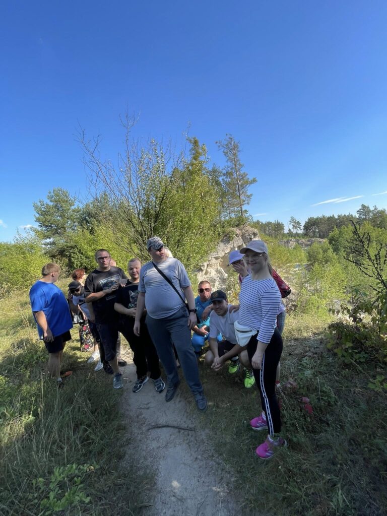 Grupa osób na szlaku turystycznym w słoneczny dzień, otoczona gęstą zielenią i krzewami.