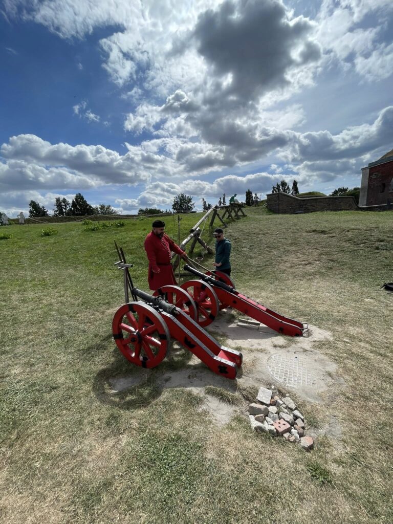 Dwóch mężczyzn w historycznych strojach przy dwóch replikach armat na trawiastym terenie zabytkowej fortyfikacji.