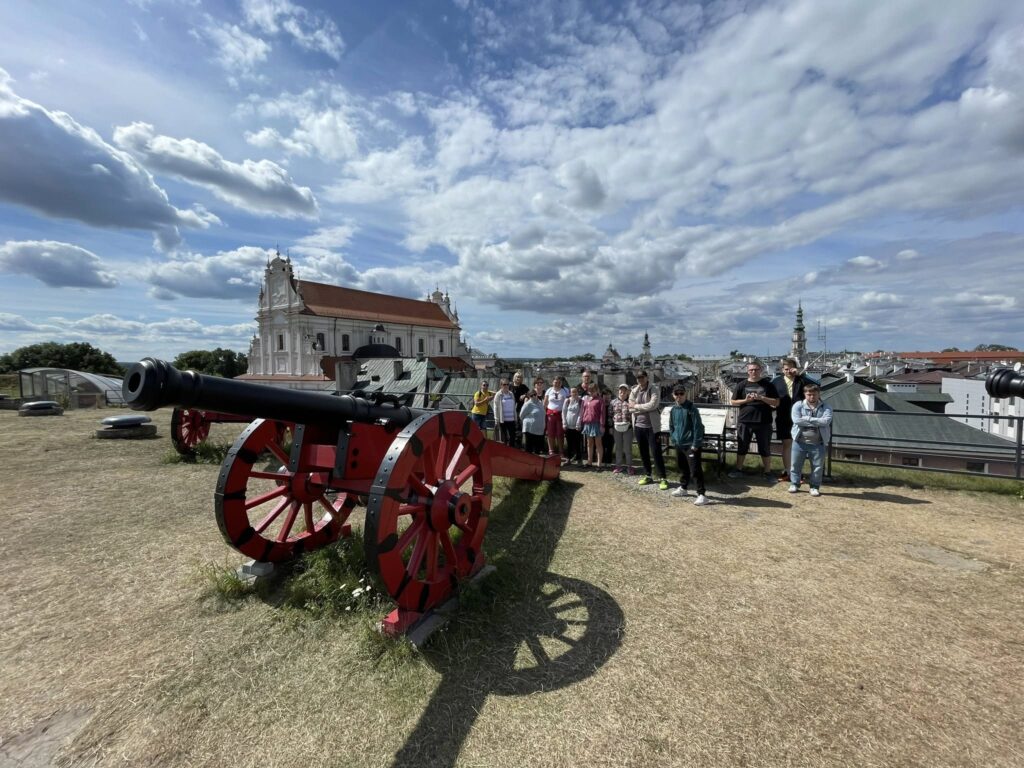 Grupa osób stojąca na murach obronnych historycznego miasta, za czerwoną armatą, z widokiem na kościół i panoramę miasta.