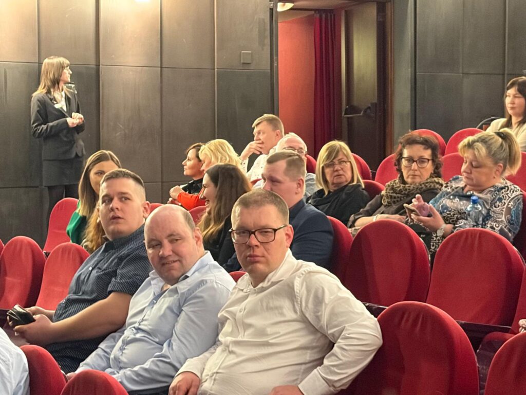 Bliskie ujęcie z widowni na grupę osób siedzących w czerwonych fotelach. Uczestnicy patrzą w stronę obiektywu, oczekując na rozpoczęcie spektaklu w sali teatralnej.