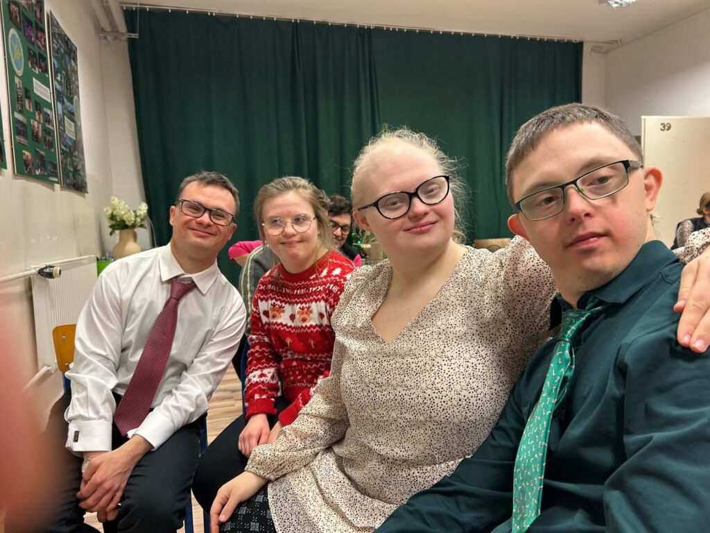 Bliskie ujęcie czwórki uśmiechniętych osób siedzących obok siebie. Uczestnicy są odświętnie ubrani, dwoje mężczyzn ma na sobie koszule i krawaty.