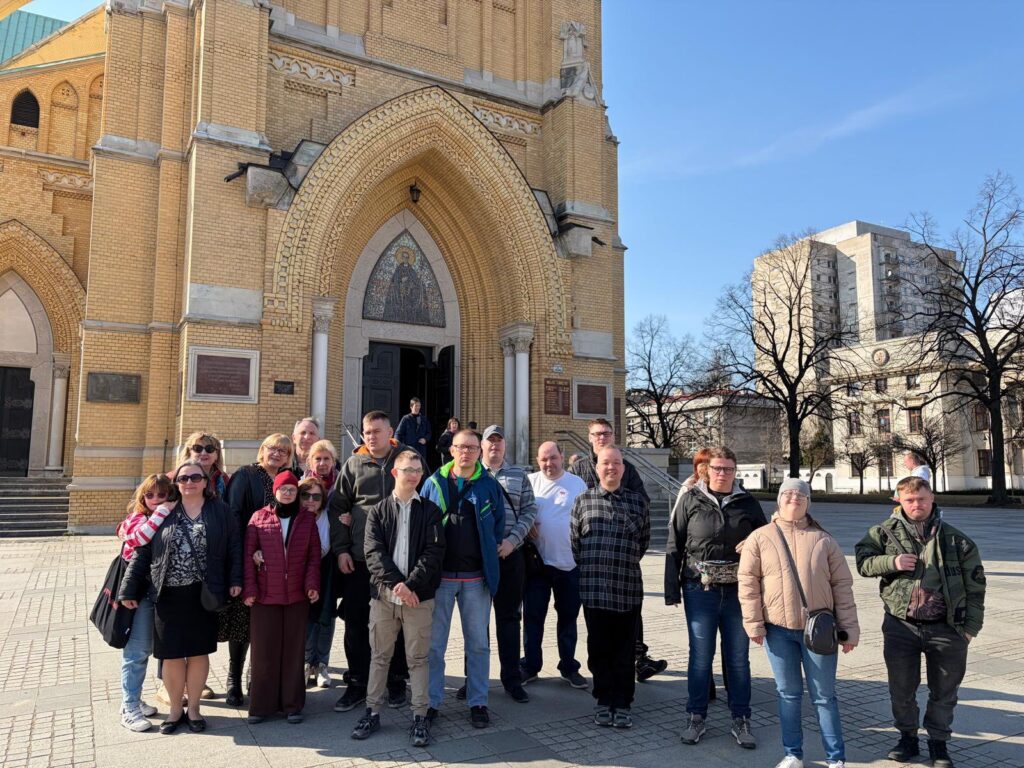 Grupowe zdjęcie uczestników wycieczki na tle wejścia do zabytkowego, ceglanego kościoła. Wszyscy stoją na placu, uśmiechnięci i zwróceni w stronę aparatu, w słoneczny, wiosenny dzień.