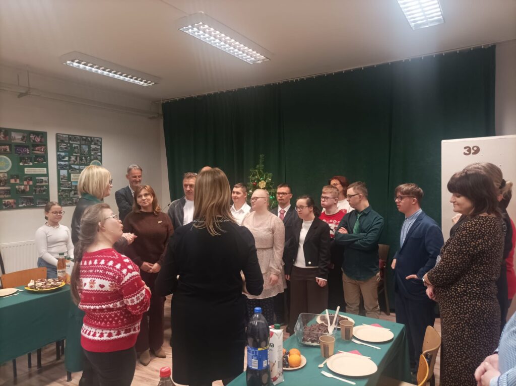 Grupa osób stoi w kręgu w jasnej sali, uczestnicząc w rozmowach i części oficjalnej spotkania. Na pierwszym planie widać stół z ciastem i napojami.