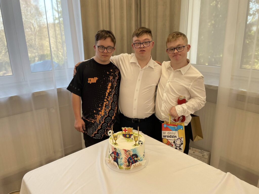 Ujęcie trzech młodych mężczyzn pozujących razem przy urodzinowym stole. Dwóch ma na sobie białe koszule, a jeden wzorzystą koszulkę. Na stole stoi tort z dekoracją piłkarską i napisami "Kuba" i "Mati".