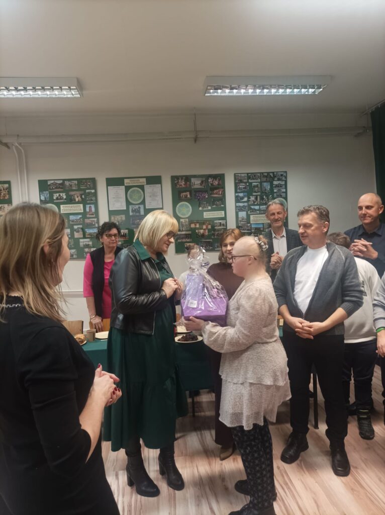 Kobieta w czarnym żakiecie wręcza młodej osobie prezent zapakowany w ozdobny, fioletowy papier. W tle grupa osób przygląda się z uśmiechem tej serdecznej chwili.