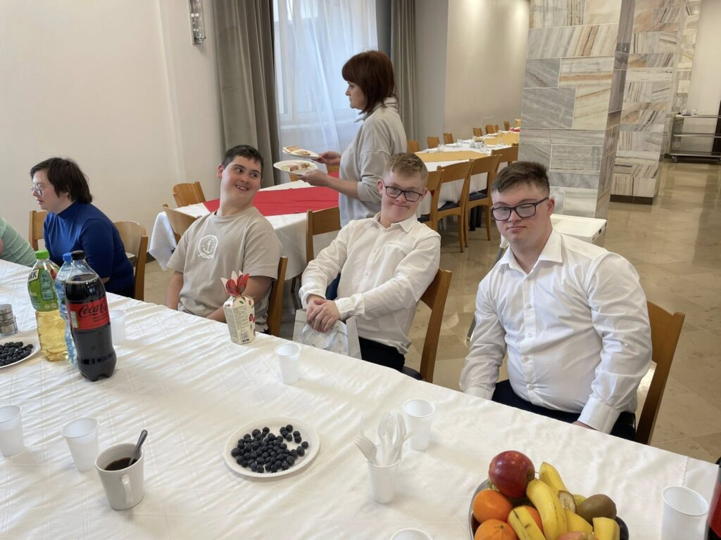 Bliskie ujęcie trzech młodych mężczyzn siedzących przy suto zastawionym biesiadnym stole. Dwóch z nich jest w białych koszulach, patrzą w stronę aparatu, uśmiechając się podczas wspólnego posiłku.