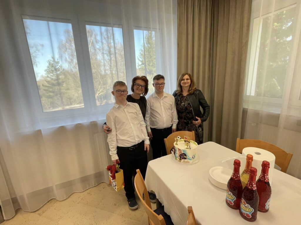 Dwóch młodych mężczyzn w białych koszulach pozuje do zdjęcia z dwiema dorosłymi kobietami. Wszyscy stoją przy stole, uśmiechnięci, w otoczeniu urodzinowych upominków i tortu.