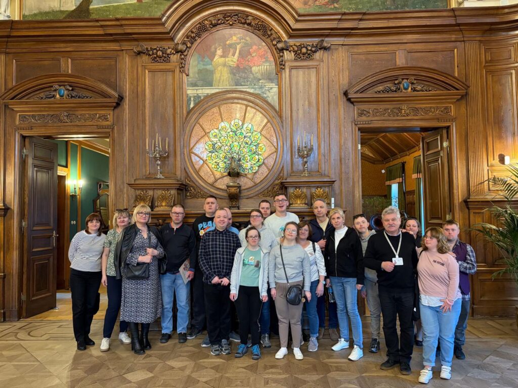 Duża grupa osób pozuje do wspólnego zdjęcia w reprezentacyjnej sali z bogatą, ciemną boazerią. Nad grupą widać ozdobny witraż z motywem pawia i malowidło ścienne.