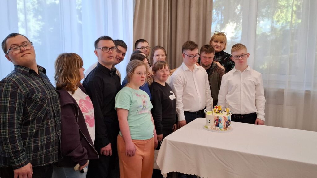 Pamiątkowe, grupowe zdjęcie uśmiechniętych młodych ludzi zebranych przy stole pokrytym białym obrusem. Na stole stoi tort urodzinowy z dekoracją piłkarską. Atmosfera jest radosna i świąteczna.