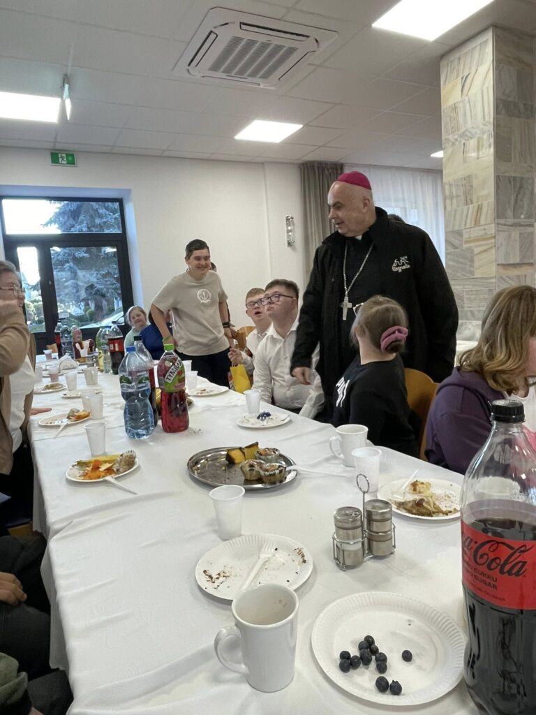 Mężczyzna w czarnej sutannie i fioletowej piusce (Biskup Adam Bab) rozmawia serdecznie z grupą młodych ludzi siedzących przy długim stole. Uczestnicy z uwagą słuchają gościa.