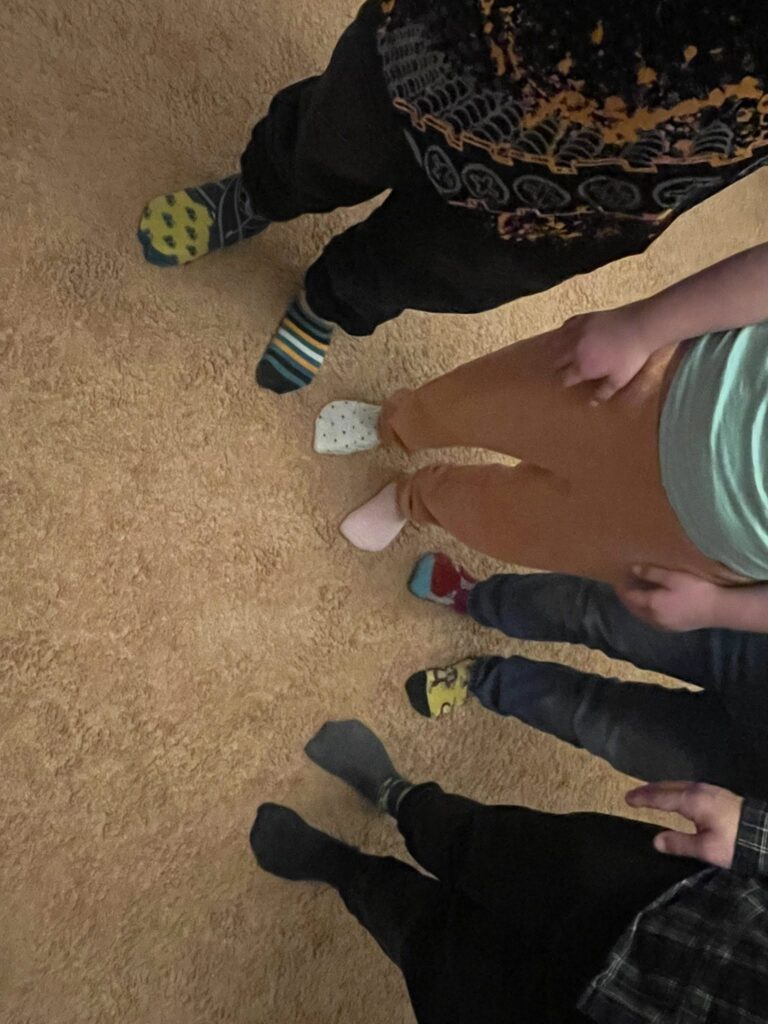 Bliskie ujęcie z góry na stopy kilku osób stojących w kręgu na puszystym dywanie. Widać różnorodne, kolorowe i wzorzyste skarpetki, co jest symbolicznym gestem w Światowym Dniu Osób z Zespołem Downa.