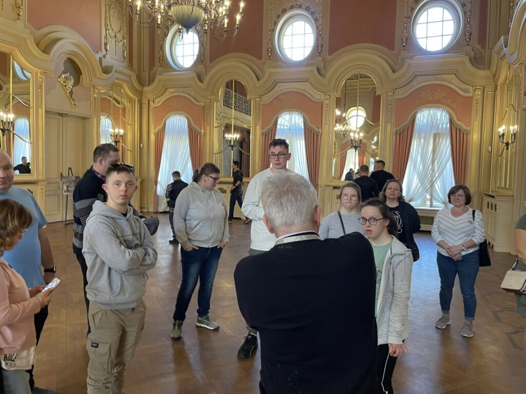 Widok na grupę zwiedzających w luksusowej sali balowej. Na pierwszym planie widać starszego mężczyznę, przewodnika, stojącego tyłem, który przemawia do zgromadzonych wokół niego osób. Wszyscy słuchają z uwagą.