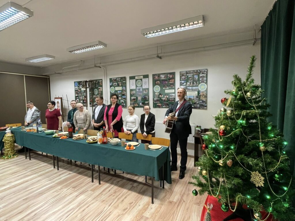 Szerokie ujęcie sali z ubraną choinką na pierwszym planie. Przy długim stole stoi grupa osób, a mężczyzna obok nich gra na gitarze, prowadząc wspólny śpiew.