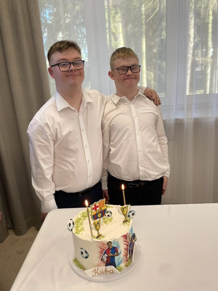 Ujęcie dwóch młodych mężczyzn w białych koszulach (Kuba i Mateusz) stojących obok siebie i z zaangażowaniem zdmuchujących płonącą świeczkę na urodzinowym torcie.