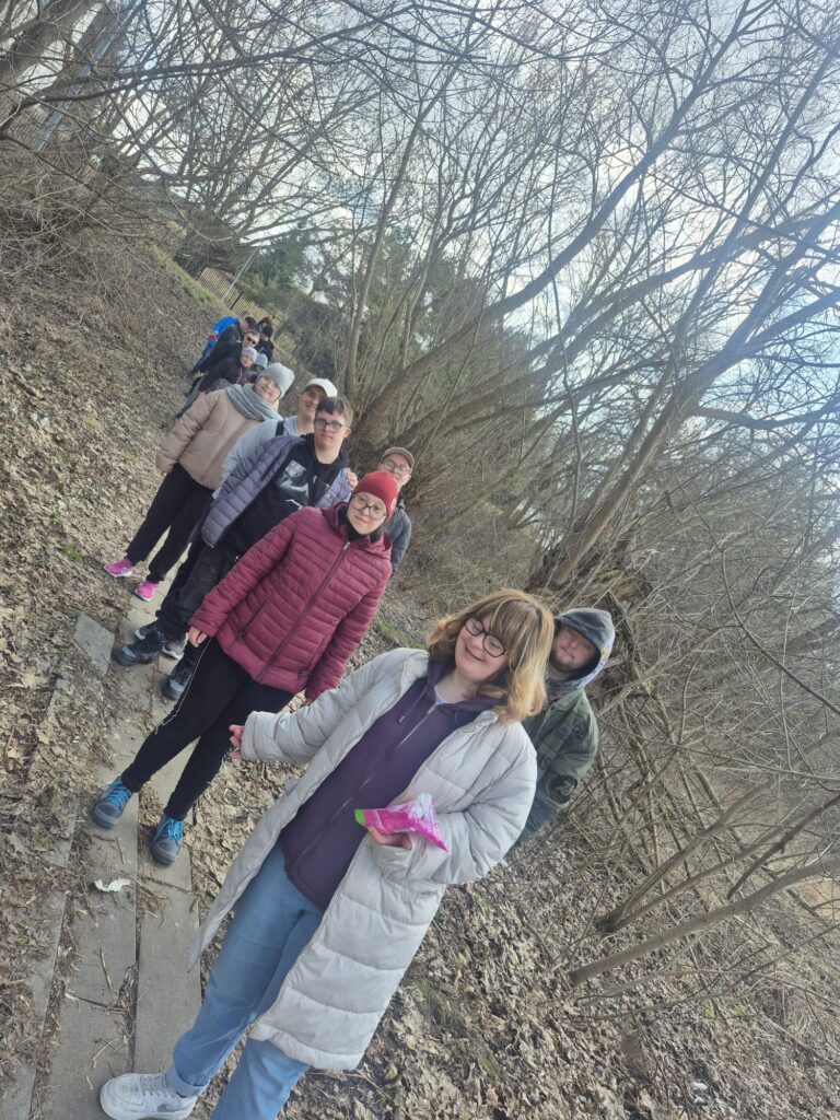 Spojrzenie z góry na grupę osób stojących w kręgu w lesie, na ścieżce pokrytej suchymi liśćmi. Uczestnicy patrzą w dół, słuchając kogoś lub obserwując coś na ziemi podczas warsztatów plenerowych.