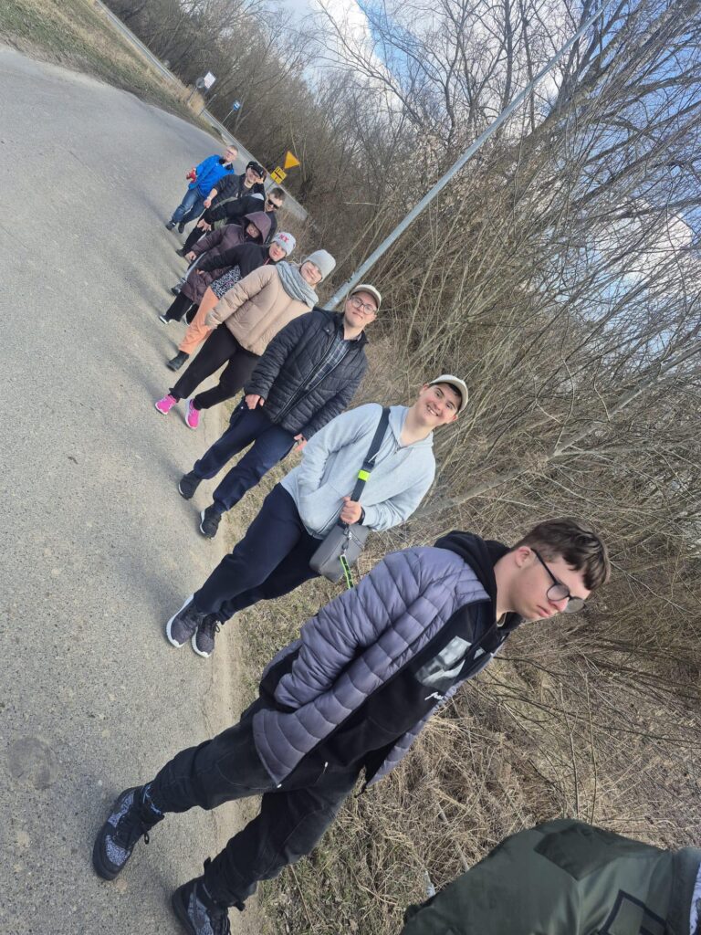 Ujęcie grupy osób idących rzędem, gęsiego, po asfalcie obok zarośli. Perspektywa z góry pokazuje dynamiczny ruch i radosną atmosferę spaceru.