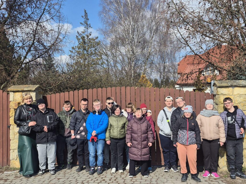 Grupowe zdjęcie kilkunastu młodych osób pozujących na tle drewnianego płotu i wejścia na posesję. Wszyscy są uśmiechnięci, ubrani w kurtki i czapki, gotowi na wspólny czas.