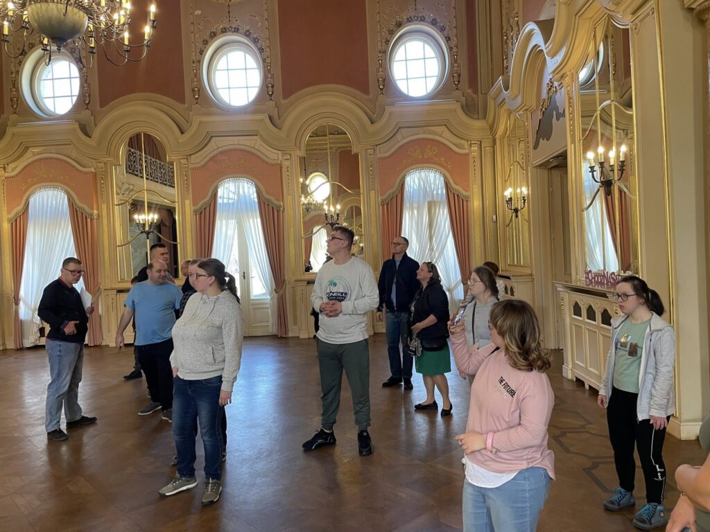 Grupa osób zwiedzających pałac stoi pośrodku wielkiej, bogato zdobionej sali balowej. Wysokie lustra i pozłacane sztukaterie odbijają światło wpadające przez okna. Uczestnicy z podziwem przyglądają się wnętrzu.