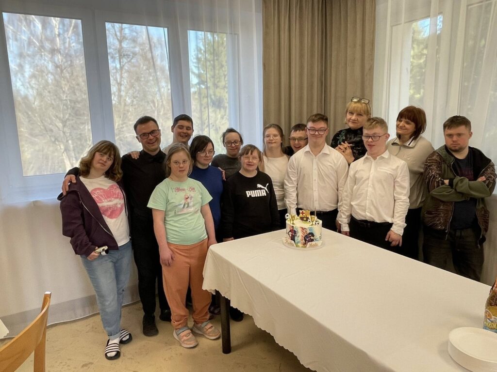 Grupowe zdjęcie kilkunastu młodych osób zgromadzonych wokół stołu urodzinowego. W centrum stoi tort, a uczestnicy patrzą w stronę aparatu, uśmiechając się podczas wspólnego świętowania.