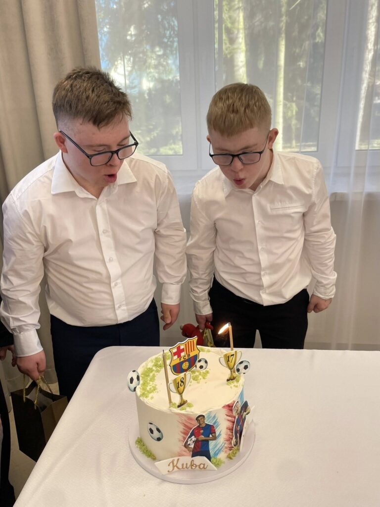 Ujęcie dwóch młodych mężczyzn w białych koszulach (Kuba i Mateusz) stojących obok siebie i z zaangażowaniem zdmuchujących płonącą świeczkę na urodzinowym torcie.