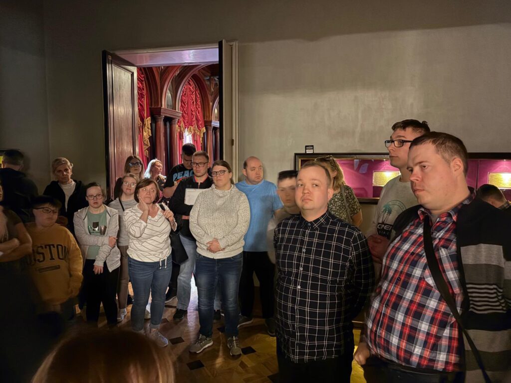 Podopieczni i opiekunowie stoją w zabytkowym, reprezentacyjnym wnętrzu, podziwiając bogate zdobienia ścian i sufitu. W tle widać wysokie okna z czerwonymi, aksamitnymi zasłonami.