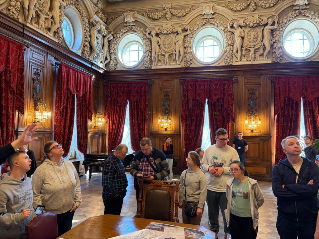 Podopieczni i opiekunowie stoją w zabytkowym, reprezentacyjnym wnętrzu, podziwiając bogate zdobienia ścian i sufitu. W tle widać wysokie okna z czerwonymi, aksamitnymi zasłonami.