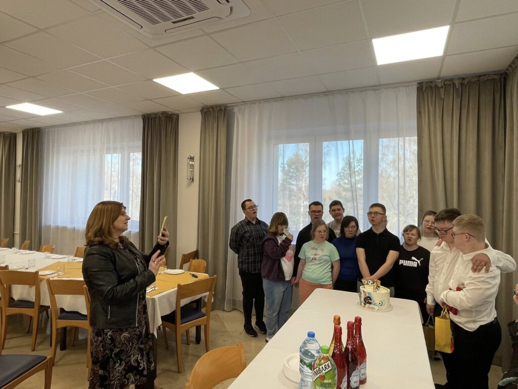 Grupowe zdjęcie kilkunastu osób (młodych i dorosłych) zgromadzonych wokół stołu urodzinowego, na którym stoi tort. Wszyscy patrzą w stronę aparatu, uśmiechając się podczas wspólnego pamiątkowego zdjęcia.