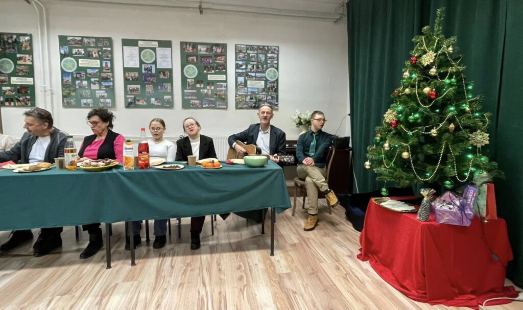Ujęcie uczestników siedzących przy stole i śpiewających wraz z mężczyzną grającym na gitarze. Po prawej stronie kadru stoi rozświetlona choinka z prezentami pod spodem.