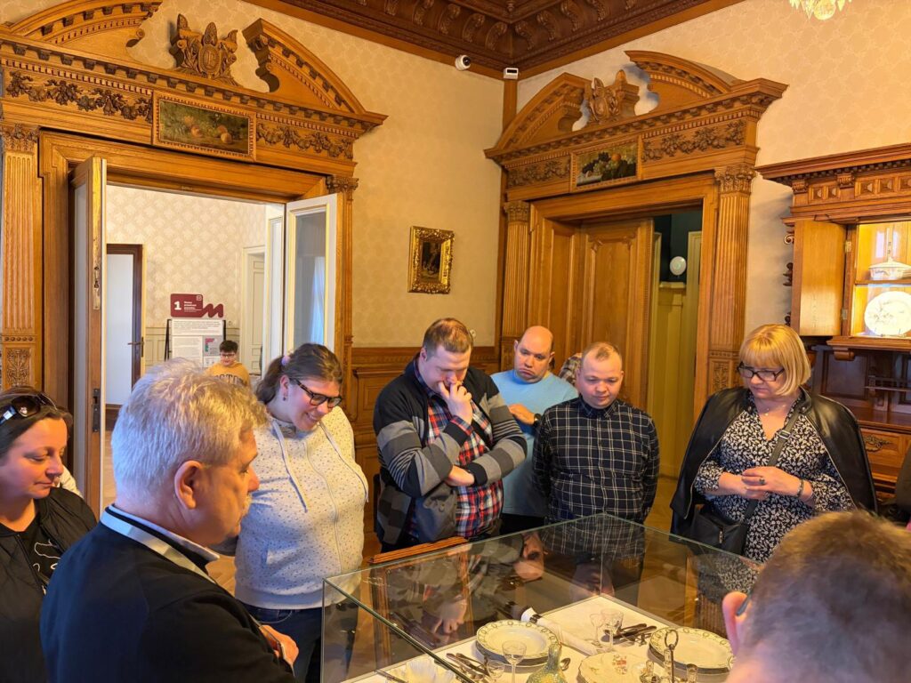 Uczestnicy wycieczki stoją wokół szklanej gabloty w zabytkowym wnętrzu z drewnianą boazerią. Z zainteresowaniem oglądają eksponowaną wewnątrz porcelanową zastawę stołową.