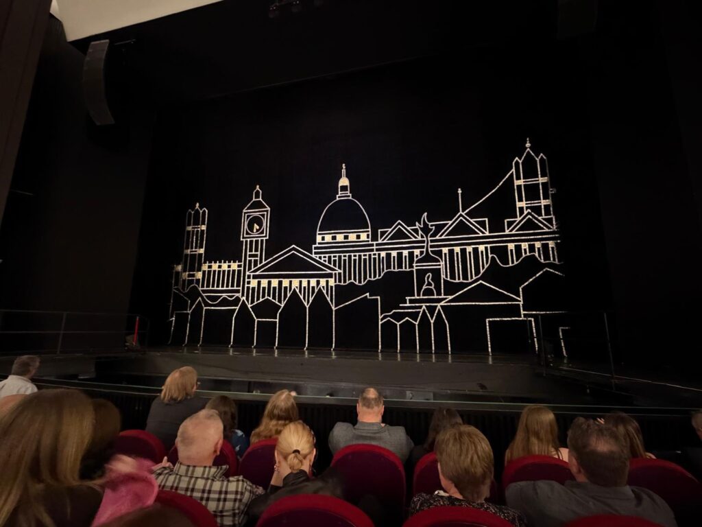 Widok z widowni na oświetloną scenę teatralną. Na czarnej kurtynie widnieje biały, konturowy rysunek panoramy miasta z zabytkowymi budynkami i mostem.