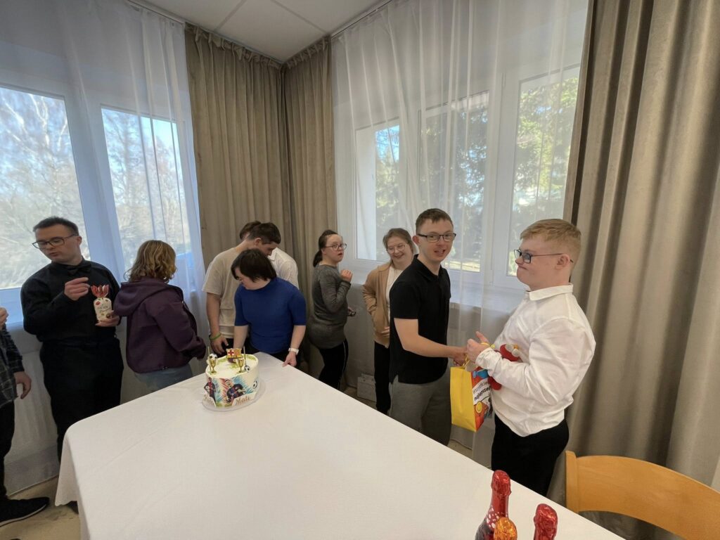 Bliskie ujęcie młodej osoby siedzącej przy urodzinowym stole. Trzyma w dłoniach upominek i tort, inne osoby stoją obok, patrząc na solenizanta z uśmiechem i gratulując.