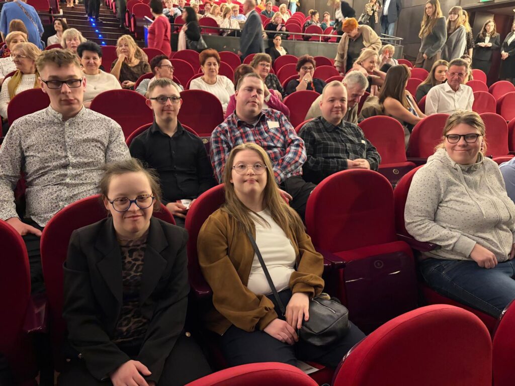 Spojrzenie na rzędy czerwonych foteli w teatrze, wypełnione publicznością. Na pierwszym planie grupa młodych osób siedzi uśmiechnięta, gotowa na obejrzenie przedstawienia.