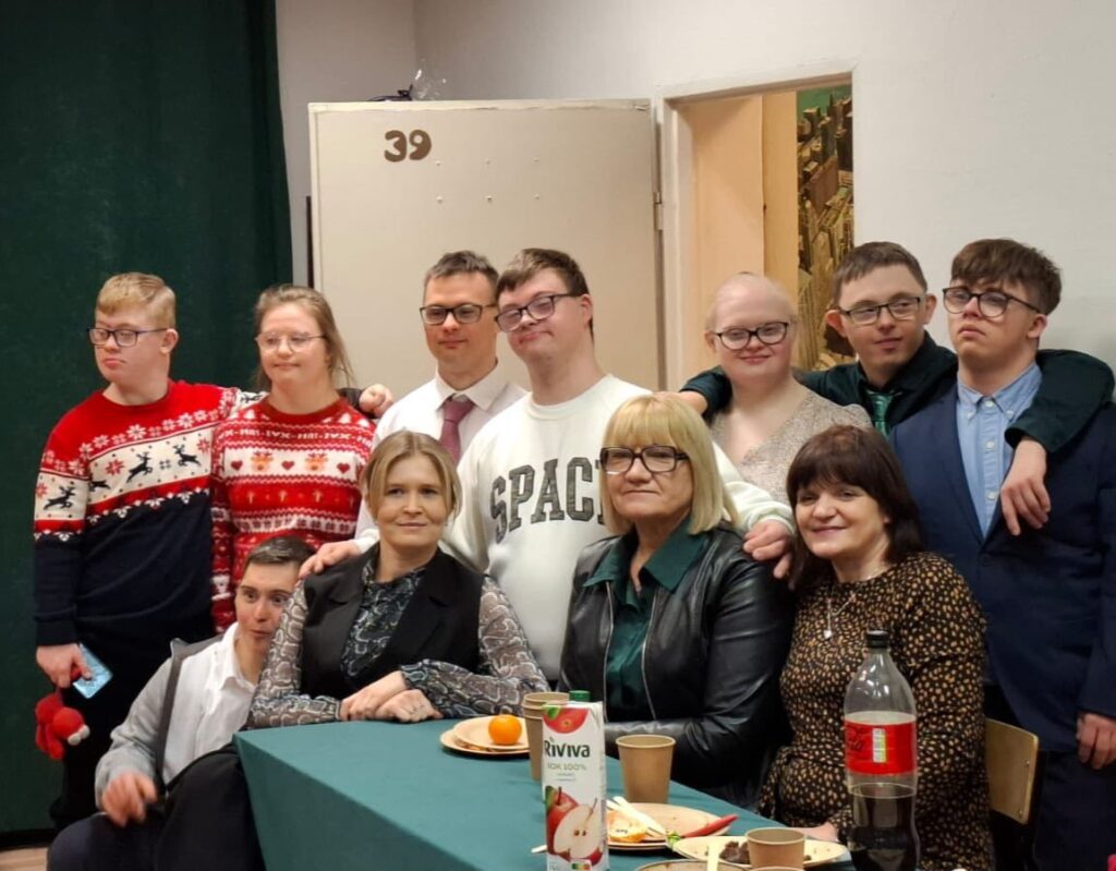 Pamiątkowe zdjęcie grupy osób zgromadzonych przy stole. Wszyscy są uśmiechnięci, niektórzy mają na sobie swetry z motywami zimowymi.