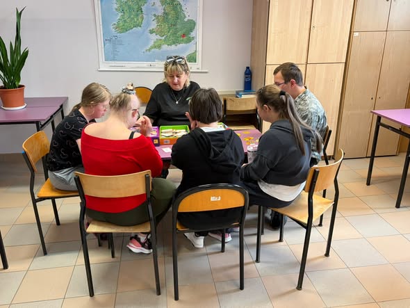 Grupa uczestników siedzi wokół fioletowego szkolnego stolika i bierze udział w grze planszowej. Dorosła opiekunka w czarnej koszulce i okularach na głowie prezentuje elementy gry z otwartego pudełka. Na ścianie w tle wisi duża mapa fizyczna Wielkiej Brytanii i Irlandii.