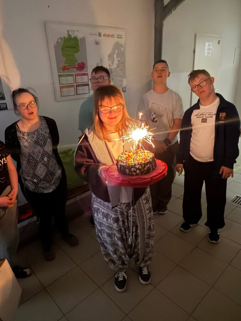 Dziewczyna w okularach i wzorzystych spodniach z uśmiechem trzyma okrągły czekoladowy tort udekorowany kolorowymi lentilkami, z którego strzela jasna iskra z zimnego ognia. Za nią stoją i klaszczą pozostali uczestnicy spotkania.