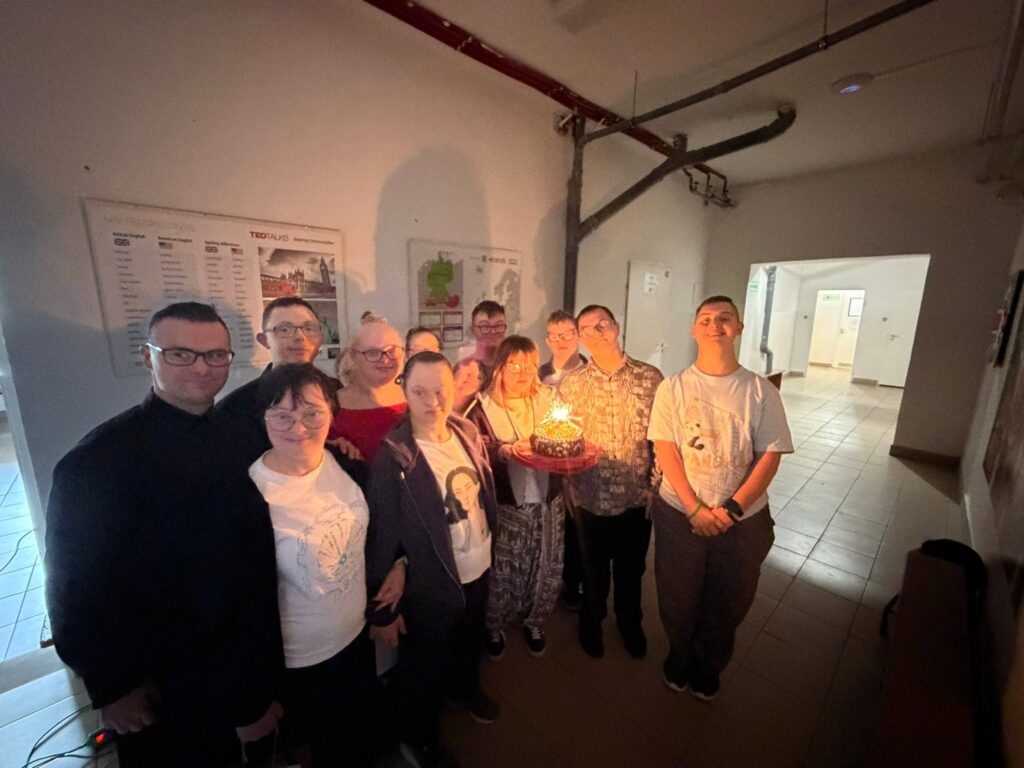 Dziewczyna w okularach i wzorzystych spodniach z uśmiechem trzyma okrągły czekoladowy tort udekorowany kolorowymi lentilkami, z którego strzela jasna iskra z zimnego ognia. Za nią stoją i klaszczą pozostali uczestnicy spotkania.