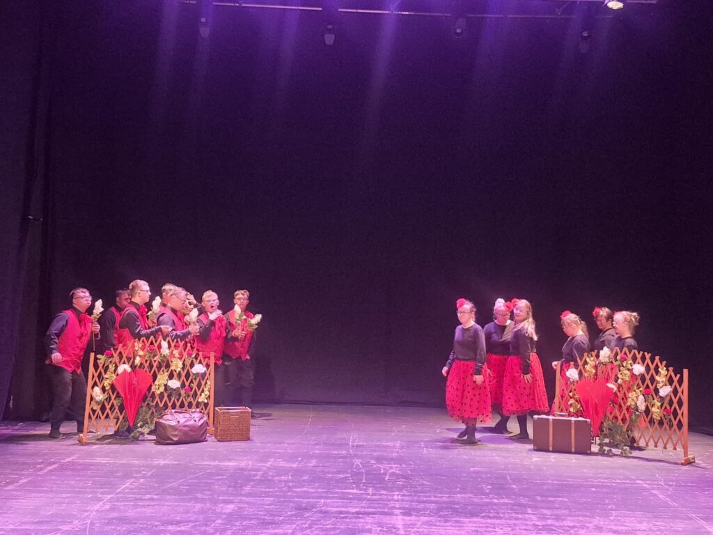 Uczestnicy przedstawienia na scenie z czerwonymi parasolkami i rekwizytami teatralnymi