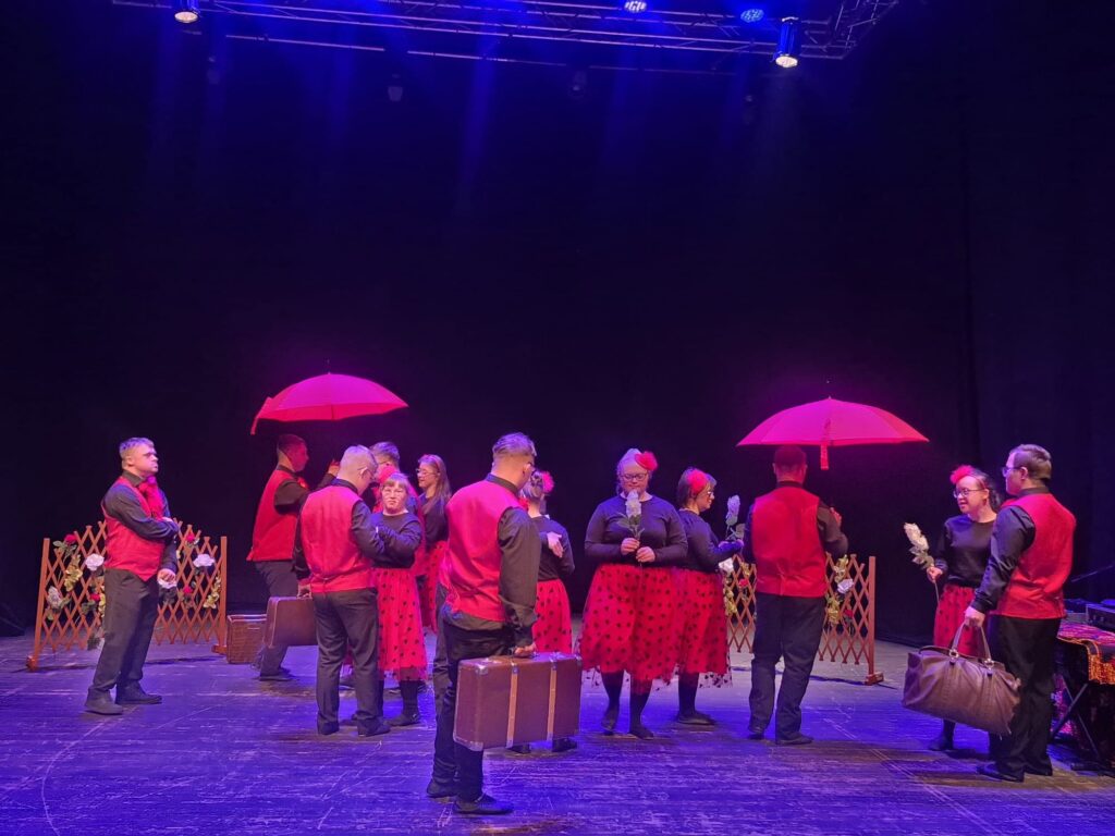 Uczestnicy przedstawienia na scenie z czerwonymi parasolkami i rekwizytami teatralnymi